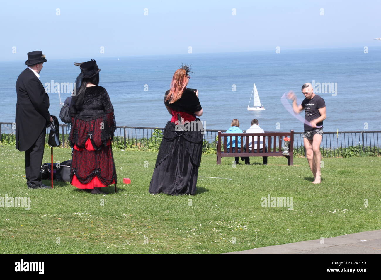 Whitby Goths watching street entertainer on the West Cliff of Whitby during Goth Weekend - beautiful blue sky and sea in the background. Stock Photo