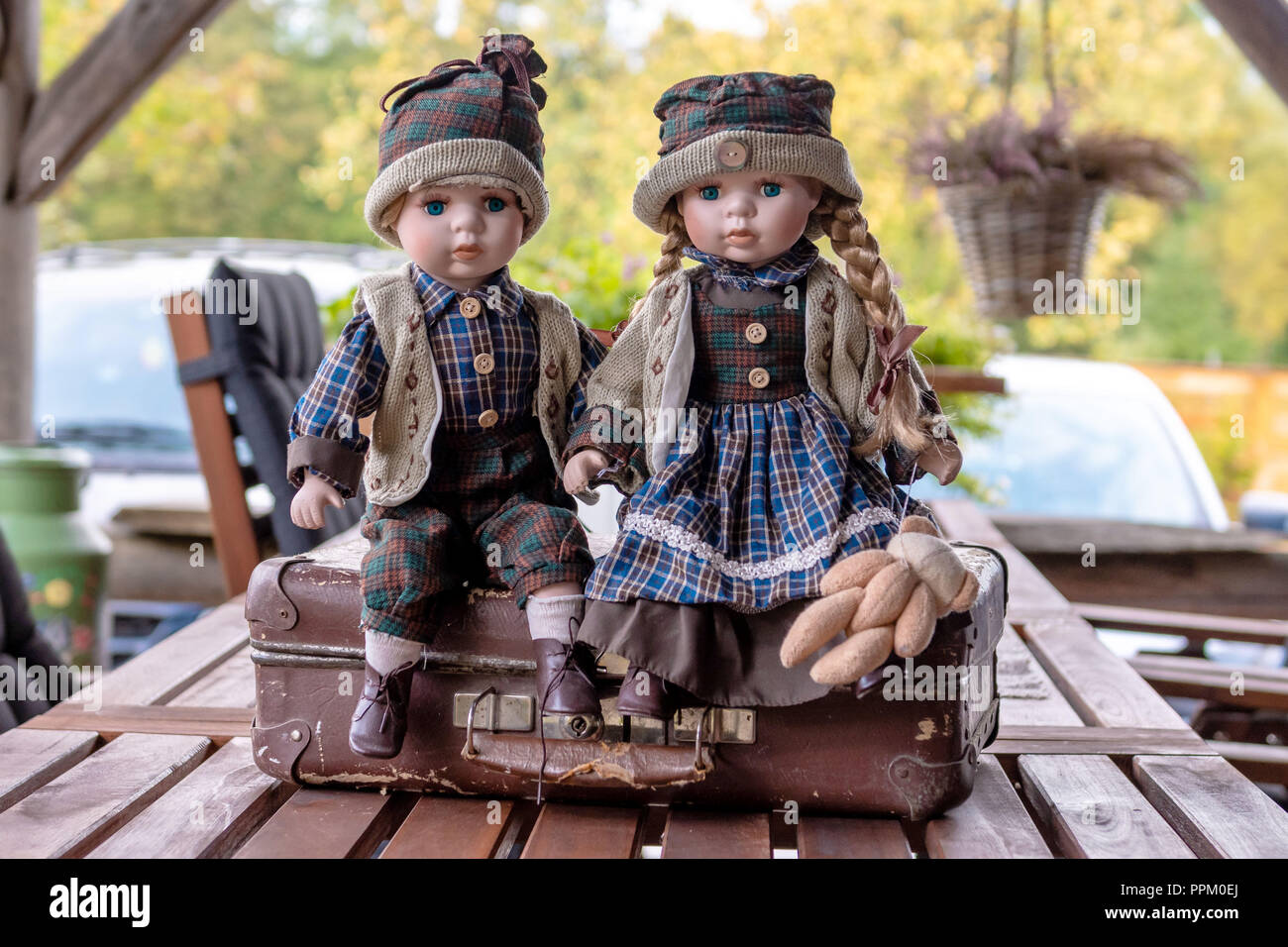 Ancient porcelain dolls sits on suitcase near log farmhouse. Stock Photo