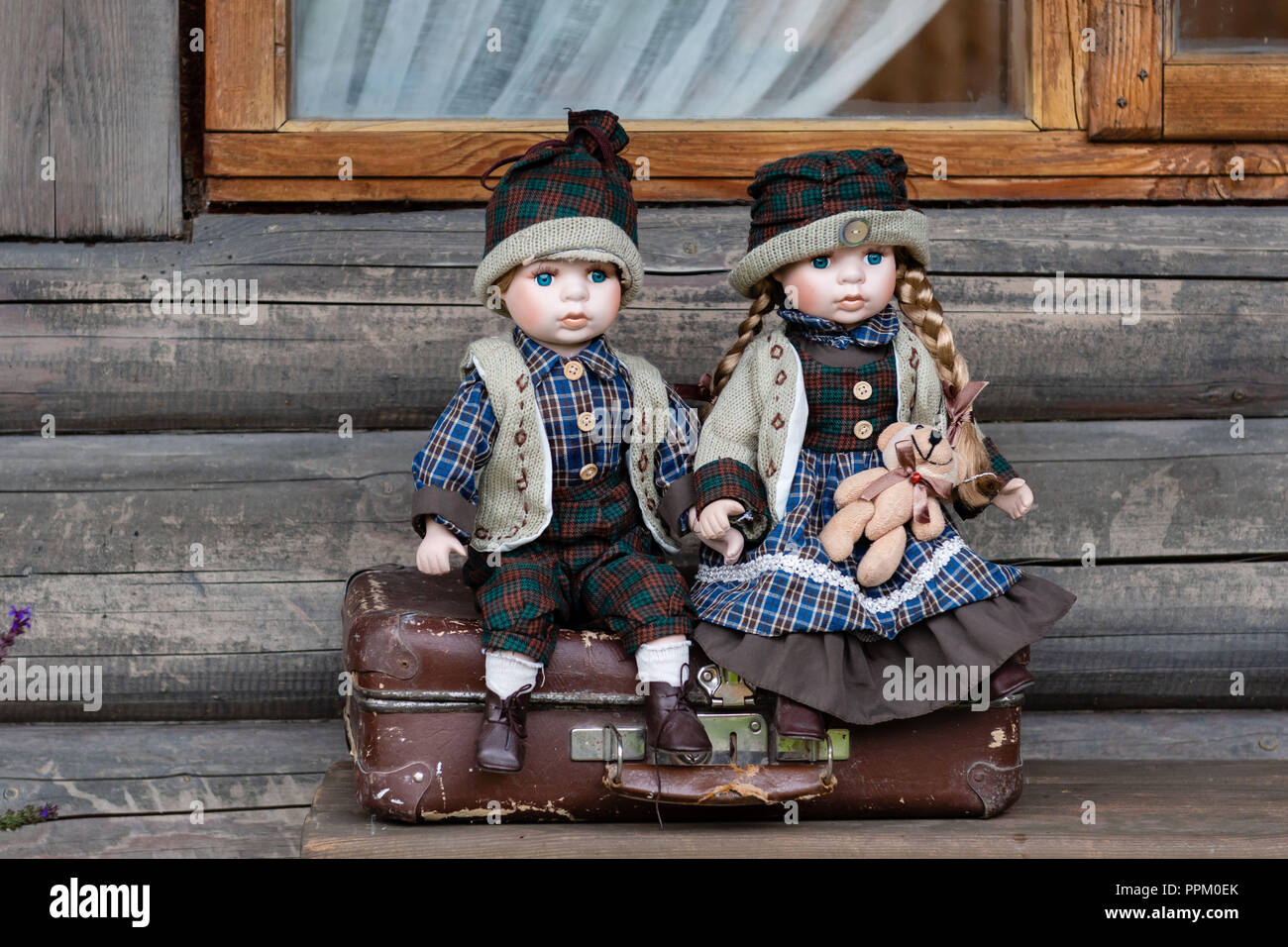 Ancient porcelain dolls sits on suitcase near log farmhouse. Stock Photo