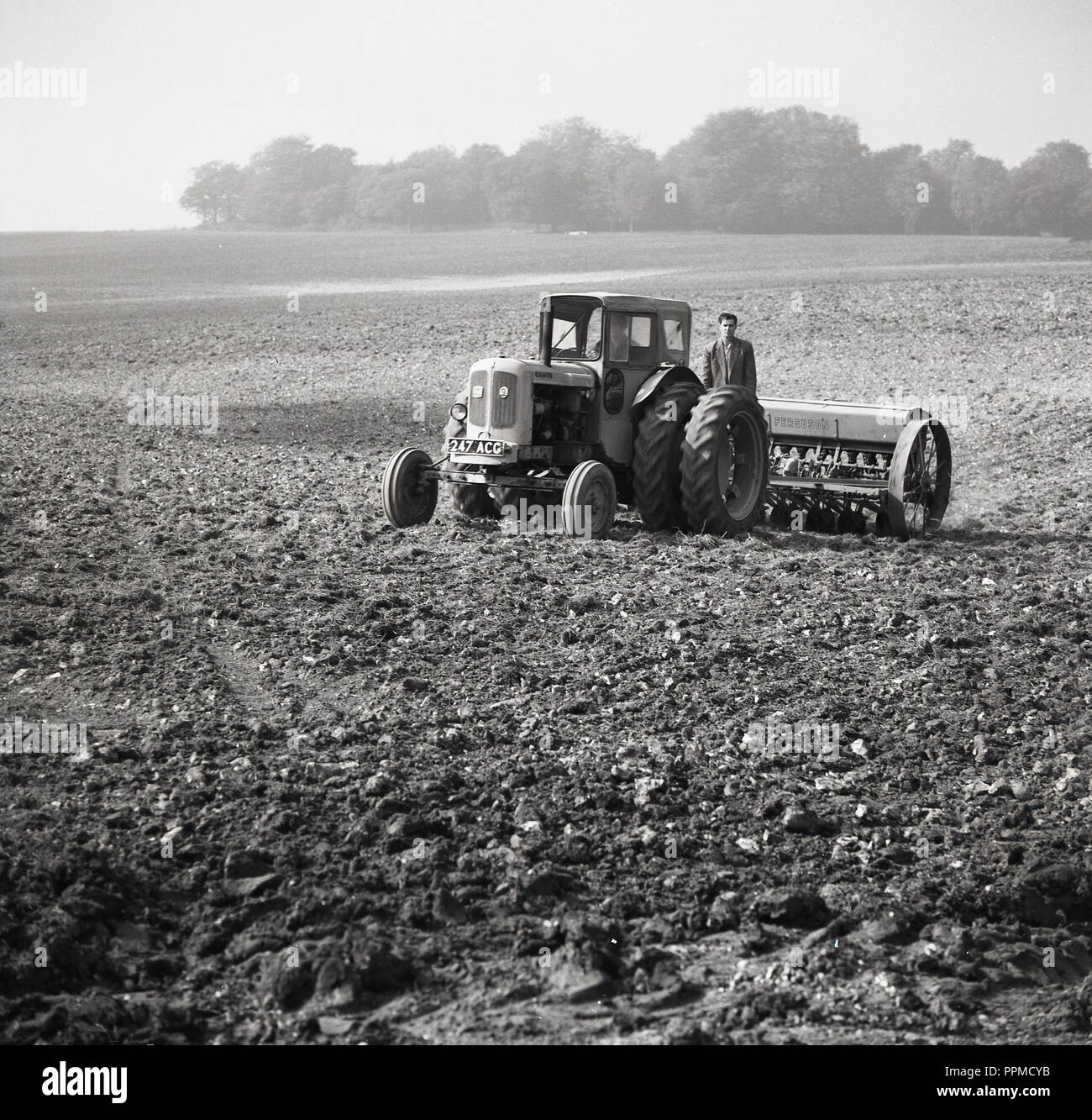 1950s, historical, in the middle of a rough ground field, a farmer stands on the back of his tractor, attached with a soil raking machine,  England, UK. Stock Photo
