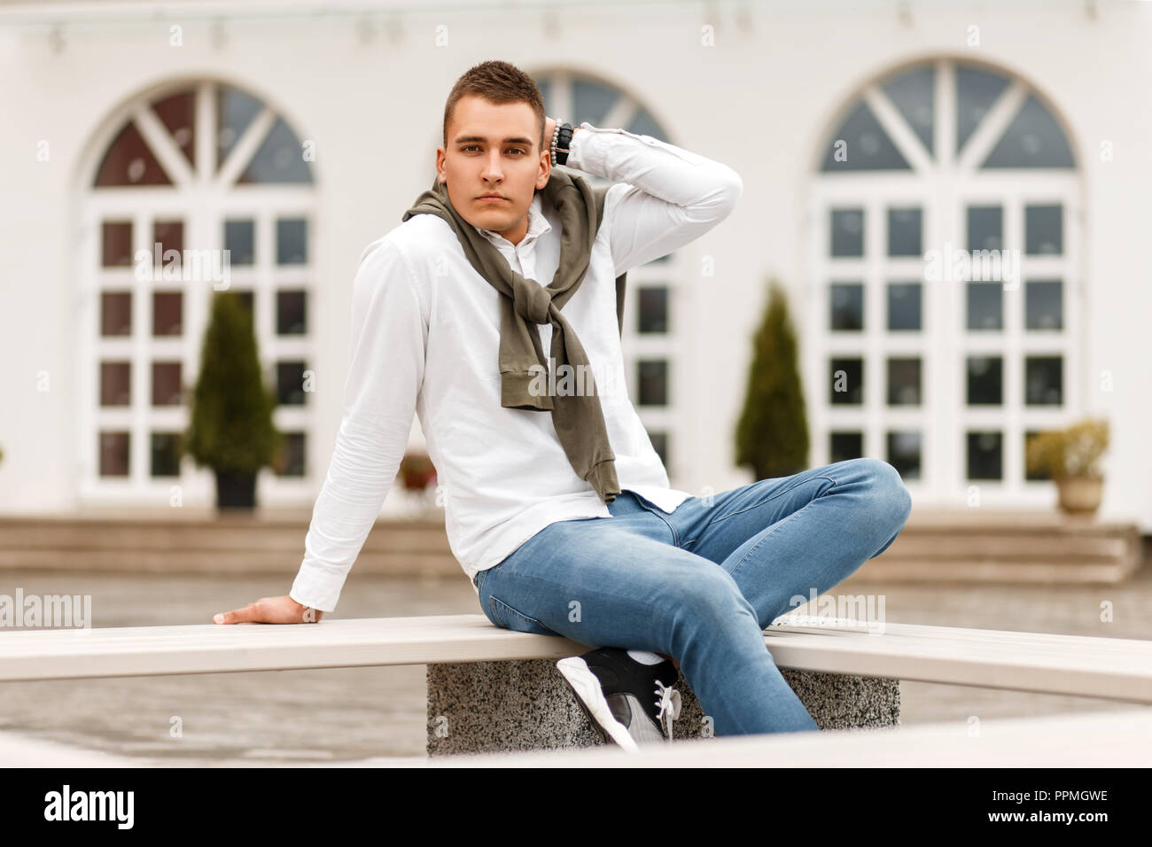 Stylish handsome young successful man in a white shirt with blue jeans sitting on a bench outside at the hotel Stock Photo