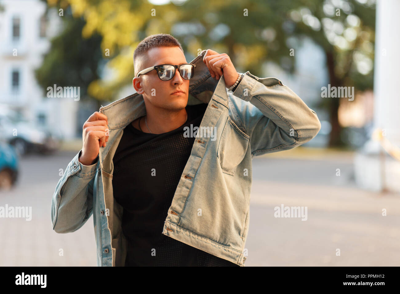 Fashionable handsome model man with a hairstyle in sunglasses and a denim jacket with a black T-shirt posing on the street Stock Photo