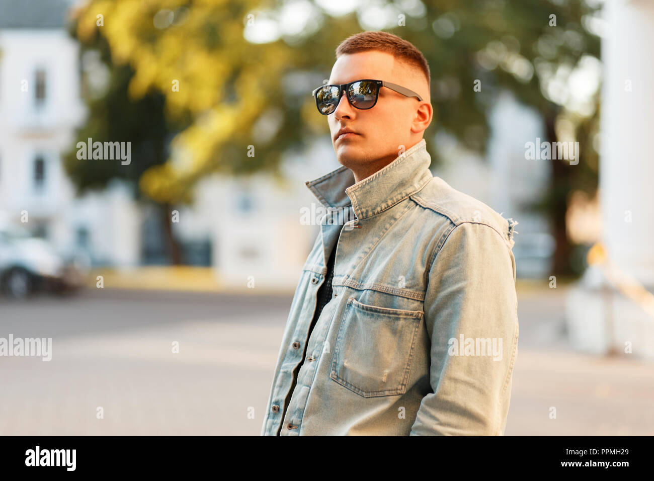 Stylish beautiful model man in a denim jacket with a black T-shirt in sunglasses in the city Stock Photo