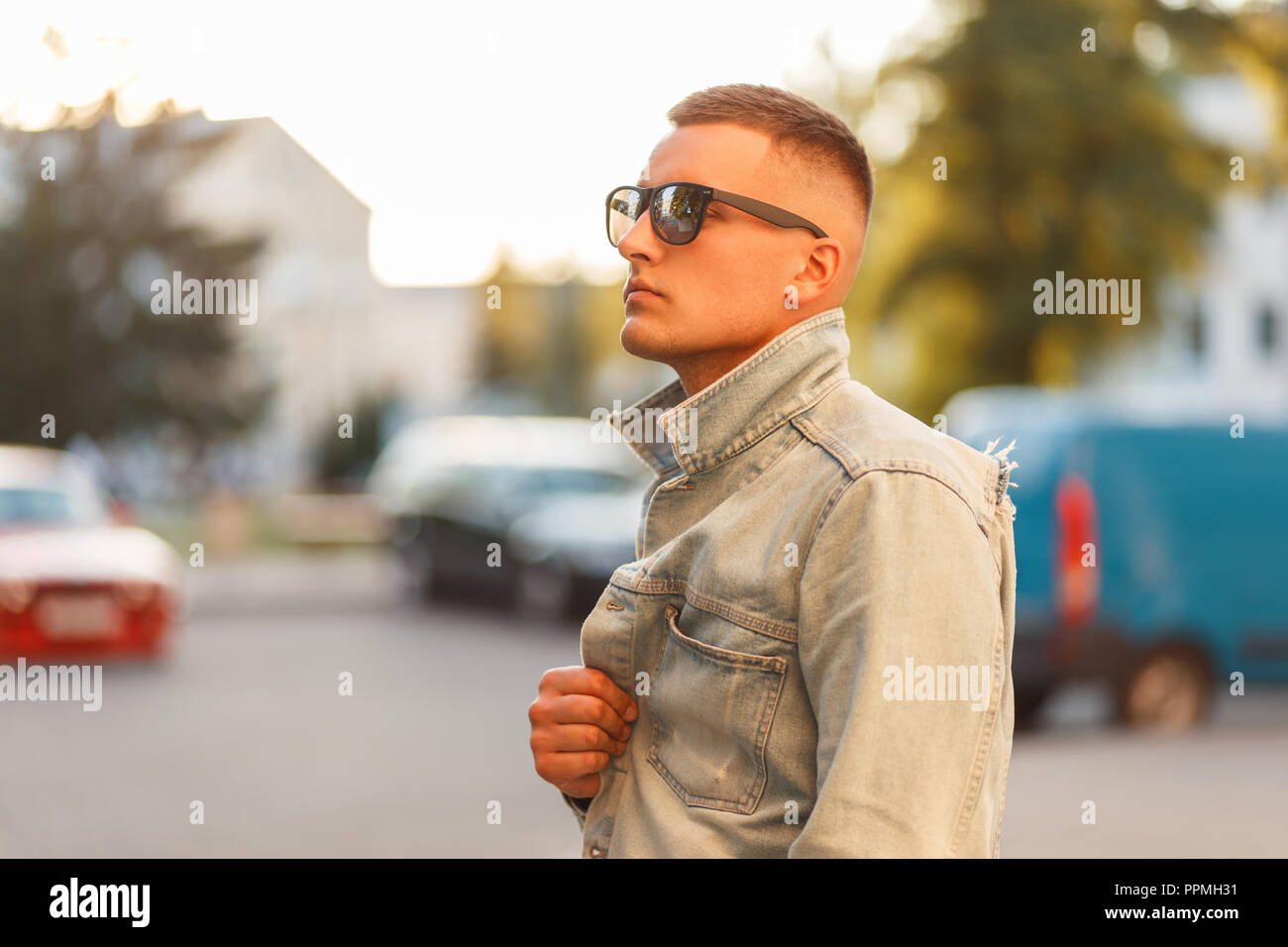 Fashionable handsome young hipster man with hairstyle in sunglasses with a black T-shirt with a jeans jacket in the street at sunset Stock Photo