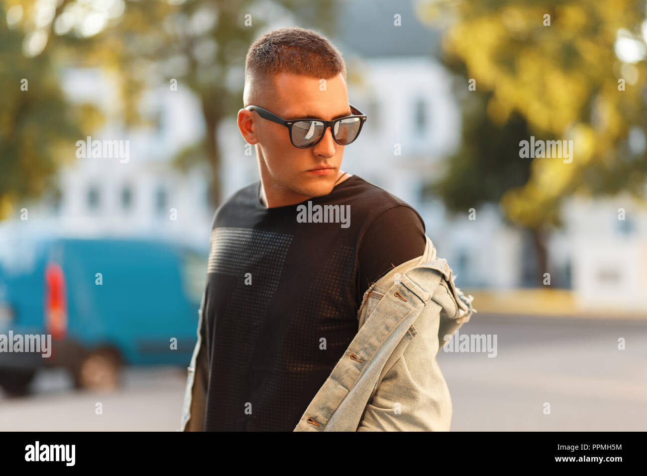 Fashionable handsome young man with fashion sunglasses in a black stylish T-shirt with a denim jacket in the city on a sunny day Stock Photo