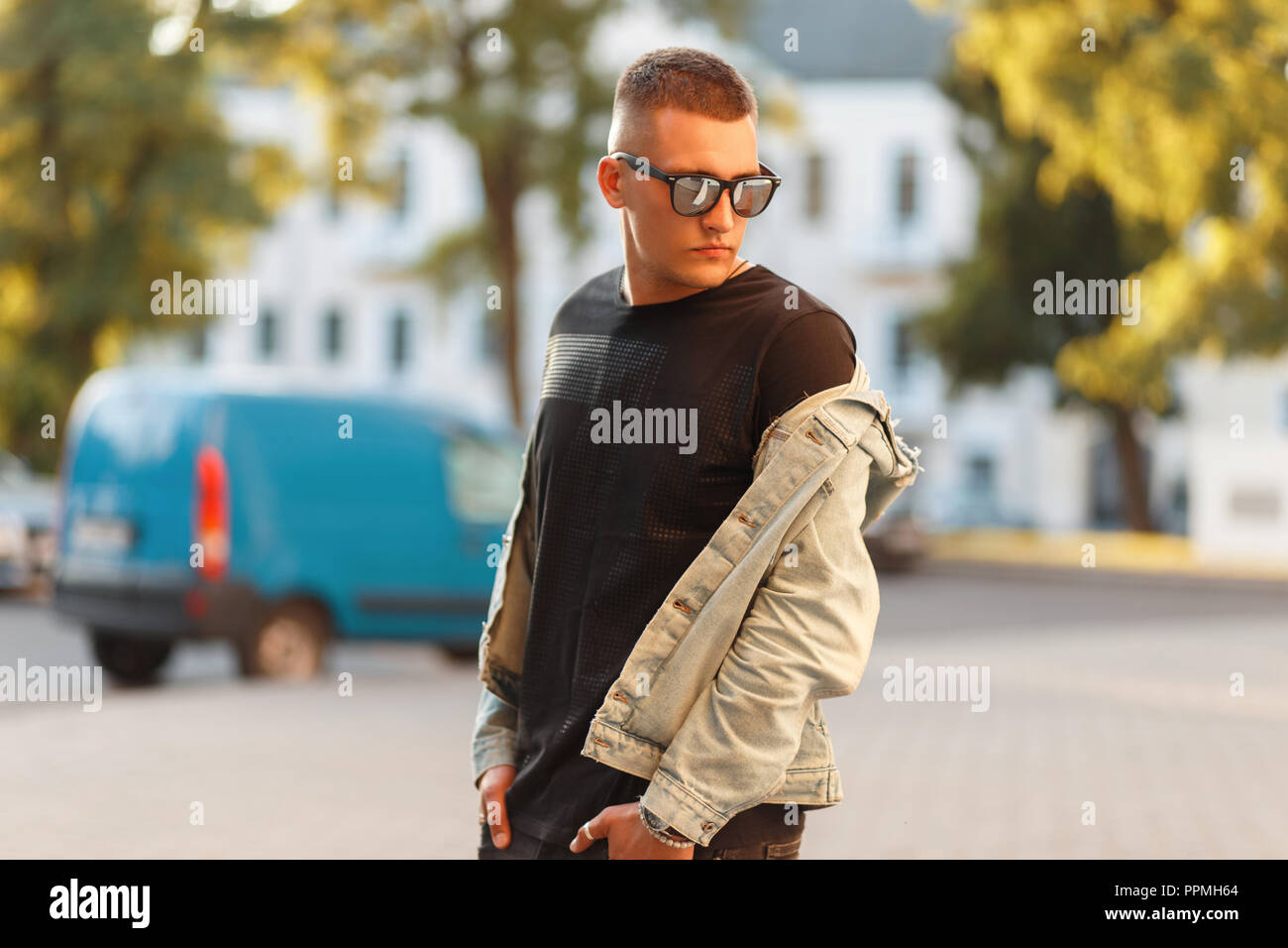 Fashionable model of a man with a hairstyle in a black stylish T-shirt with a denim jacket posing in the city Stock Photo