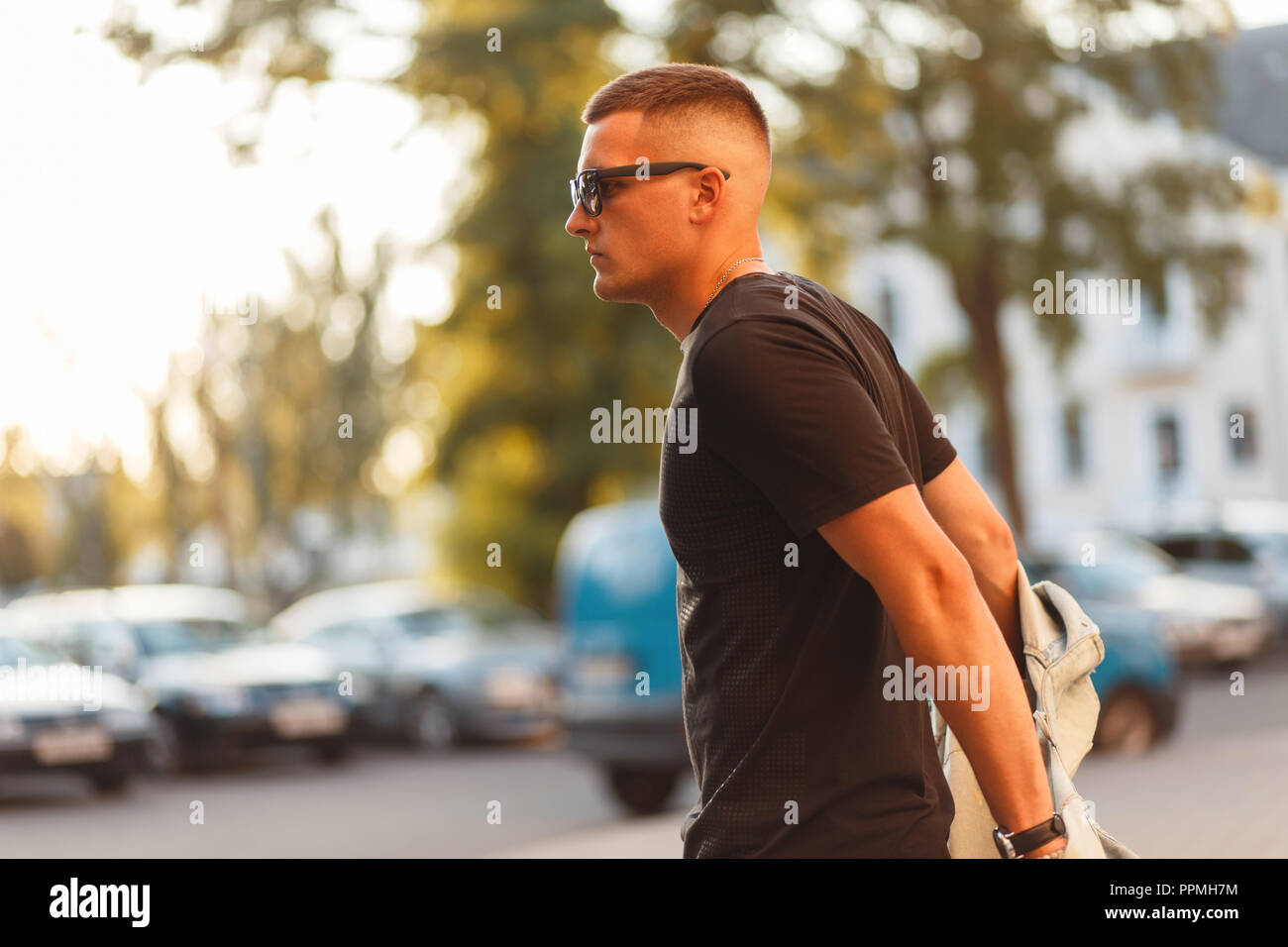 Stylish model man with a hairstyle with glasses with a black fashion T-shirt and jacket walking in the city on a sunny day Stock Photo