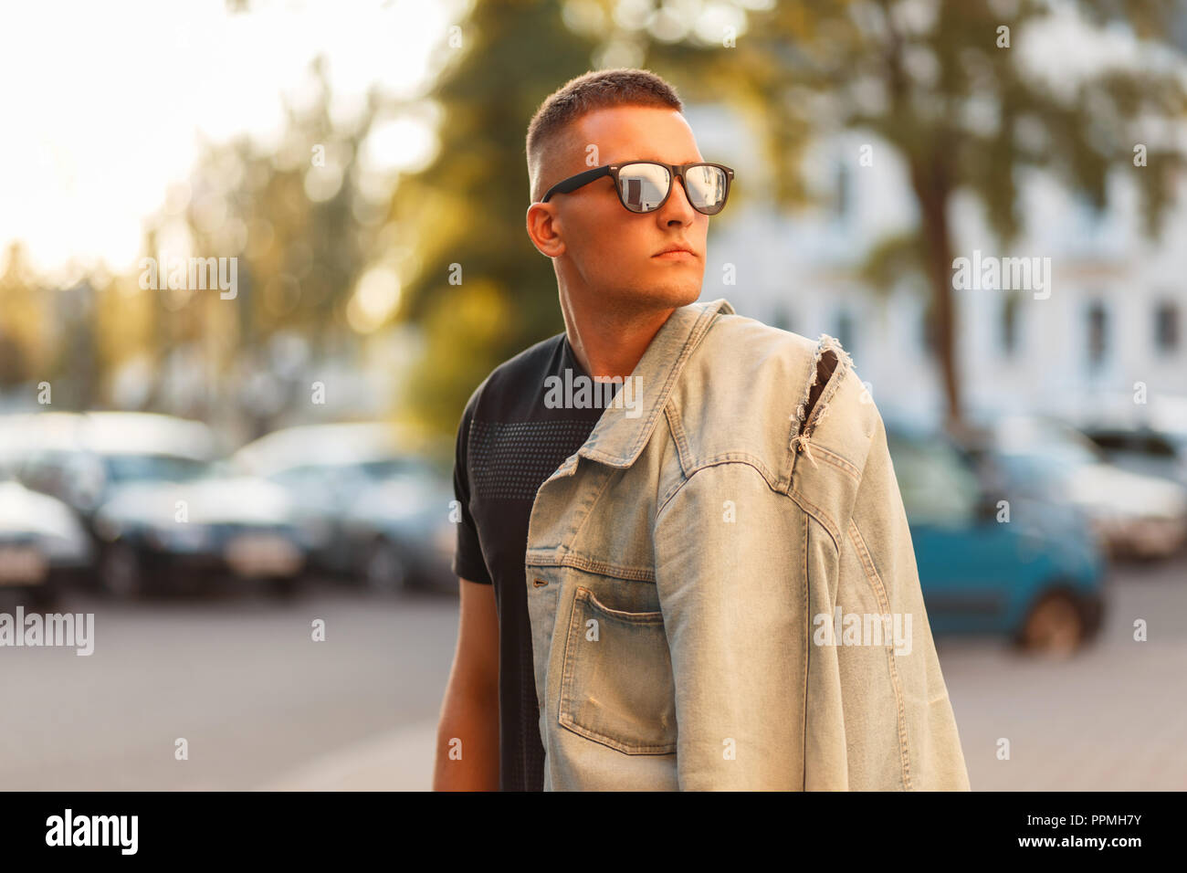 Young stylish handsome hipster man with sunglasses in a black T-shirt with a jeans jacket in the street on a sunny day Stock Photo