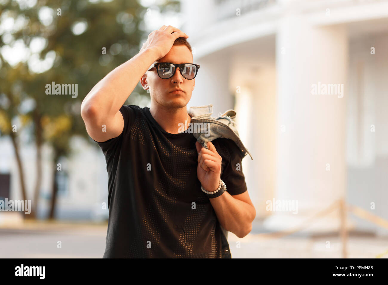 Stylish hipster man with a hairstyle in sunglasses in a black fashion T-shirt with a jacket in the city Stock Photo