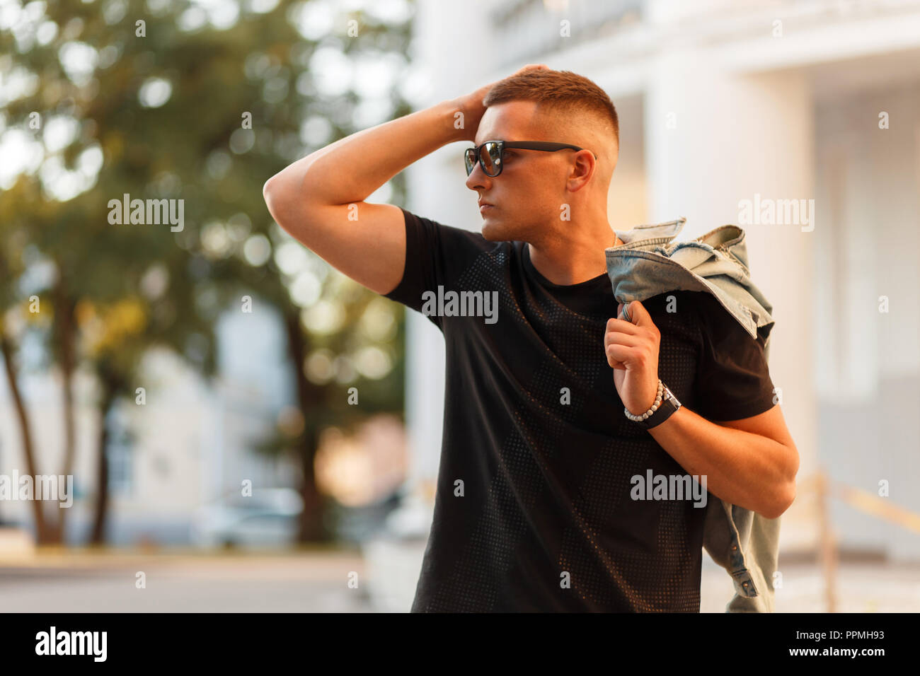 handsome fashionable hipster man with sunglasses in a black T-shirt with a jeans jacket in the street on a sunny day Stock Photo