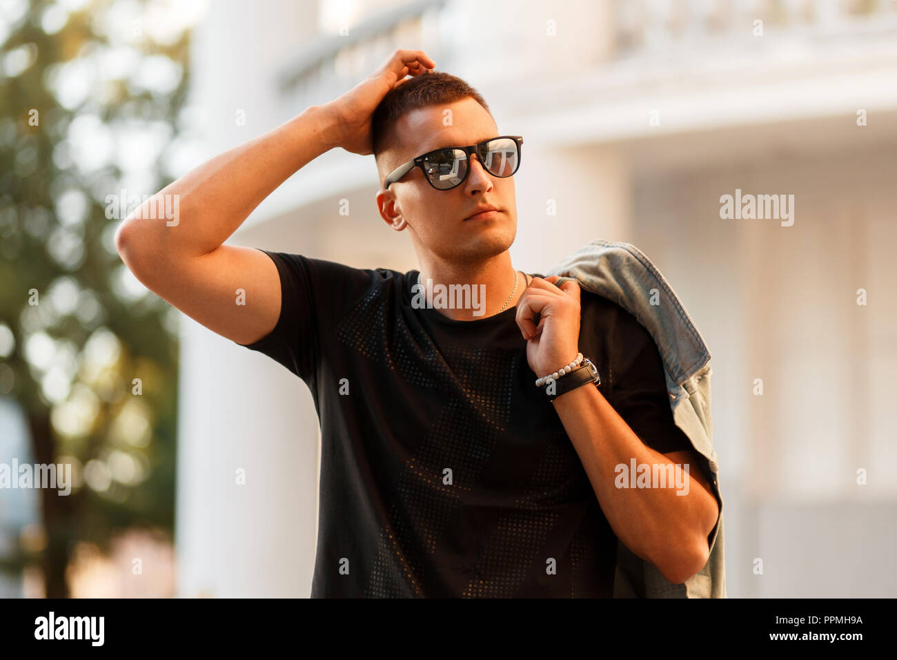 Fashionable handsome young man with a hairstyle in glasses with a black T-shirt and a denim jacket walking in the city Stock Photo