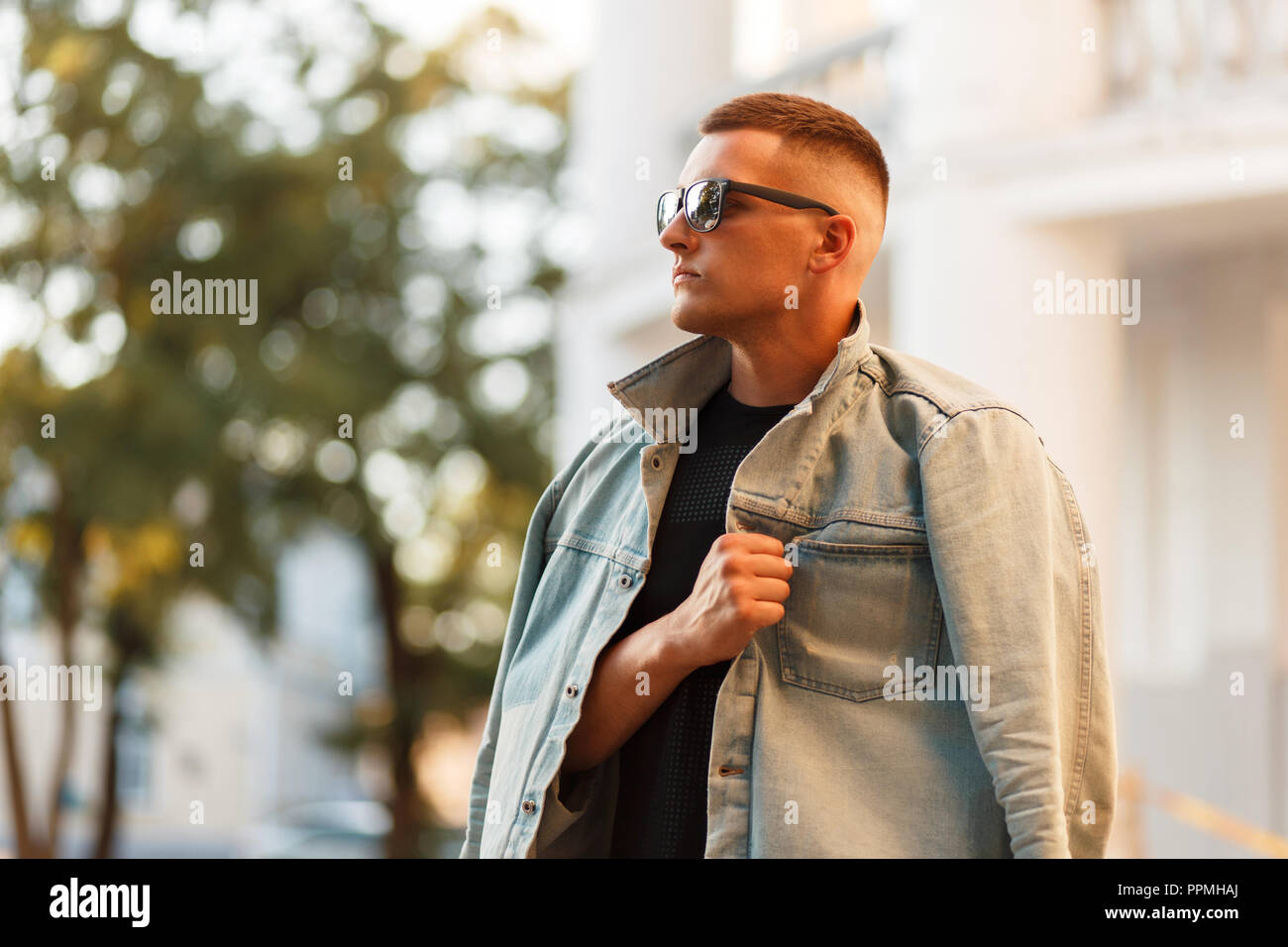 Stylish handsome young man with a hairstyle in a jeans jacket and a black T-shirt with sunglasses posing in the city Stock Photo