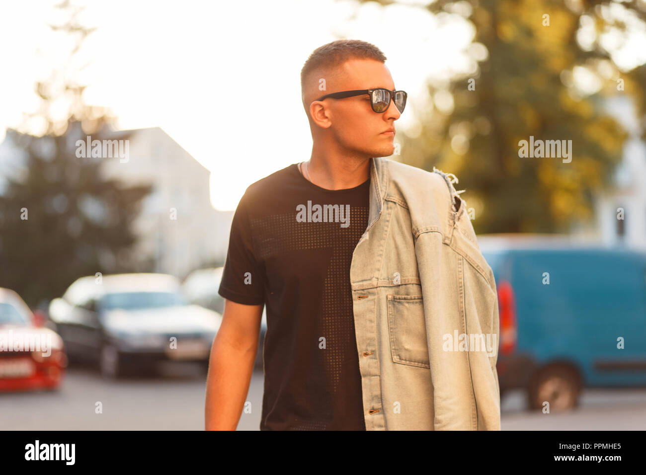 Stylish beautiful model hipster man with a hairstyle with sunglasses in a black fashion T-shirt and a vintage denim jacket walking in the city Stock Photo