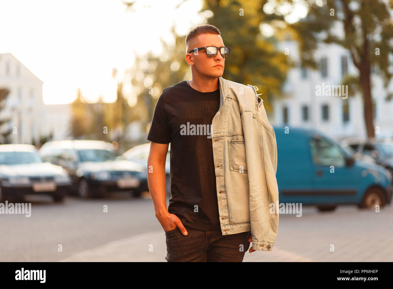Stylish handsome hipster man with a haircut in sunglasses and a fashionable black T-shirt with a denim jacket walking in the city Stock Photo