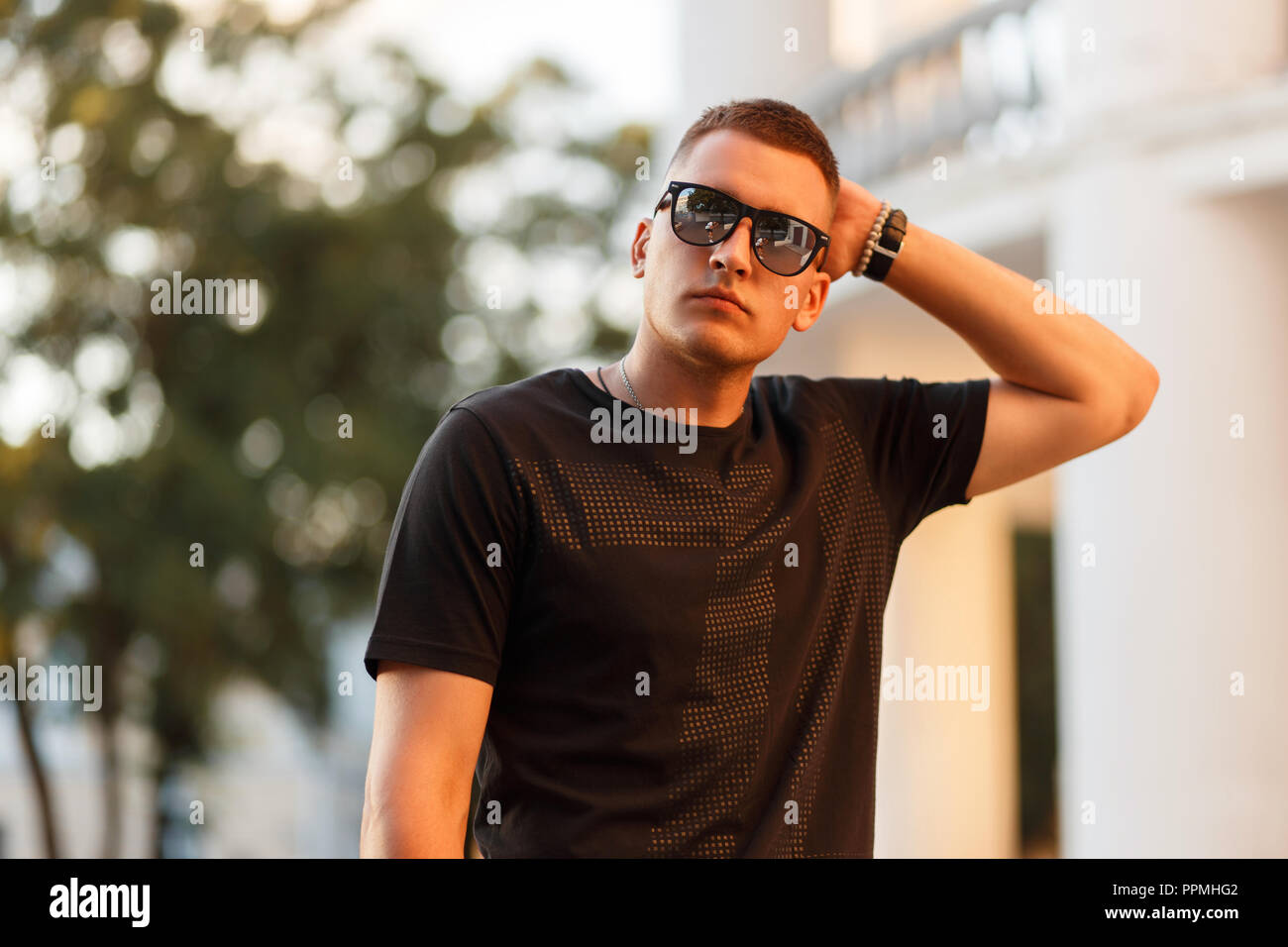 Stylish handsome man with sunglasses in a fashionable black T-shirt in the city at sunset Stock Photo