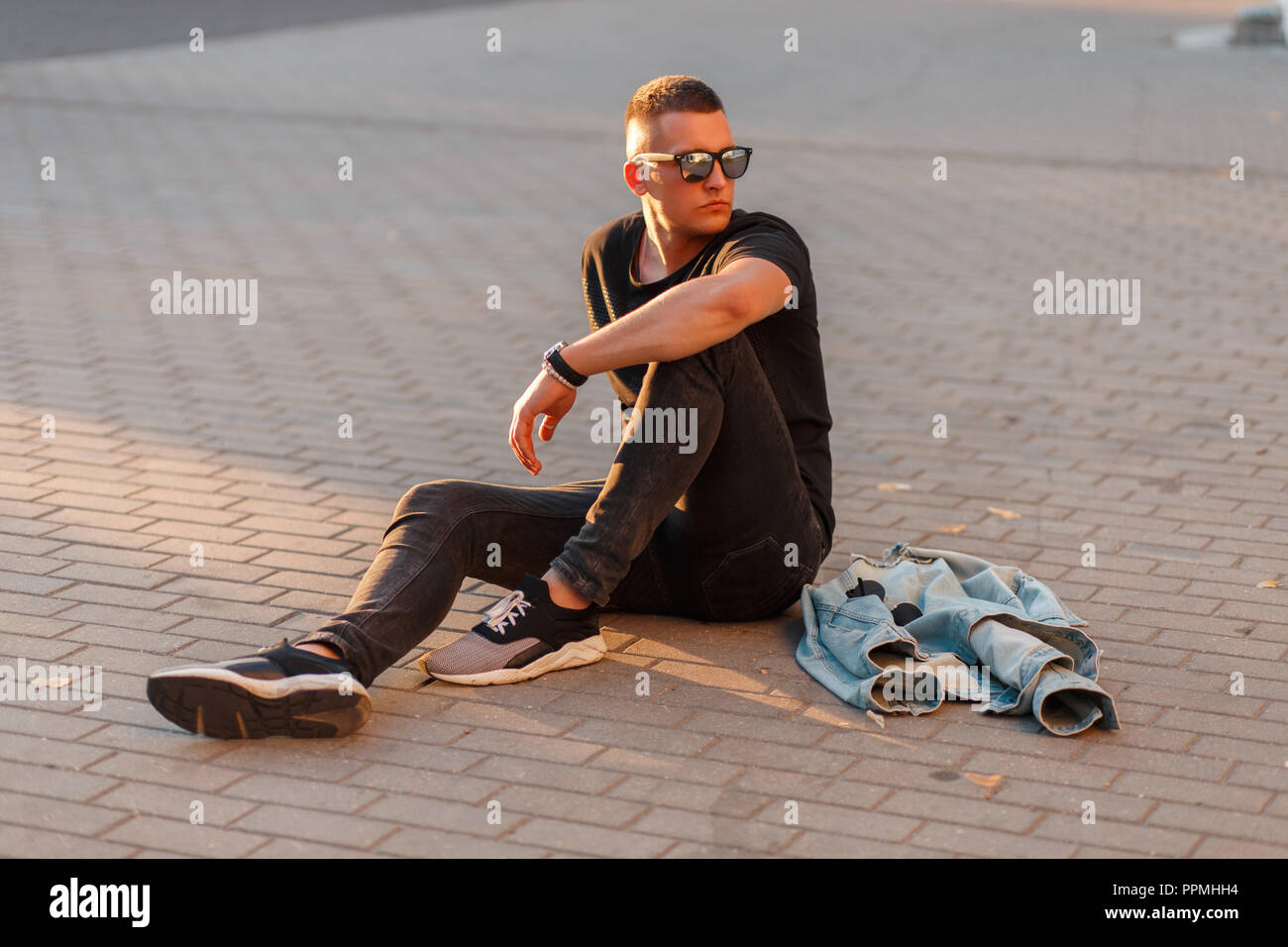 Stylish handsome young guy with glasses in black fashion clothes with sneakers sits on the street Stock Photo