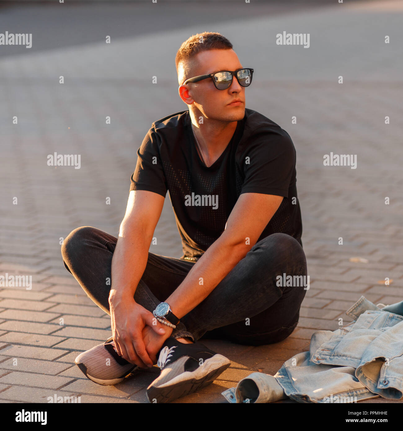 Fashionable handsome young man model in black sunglasses in a black stylish T-shirt sitting on the street Stock Photo