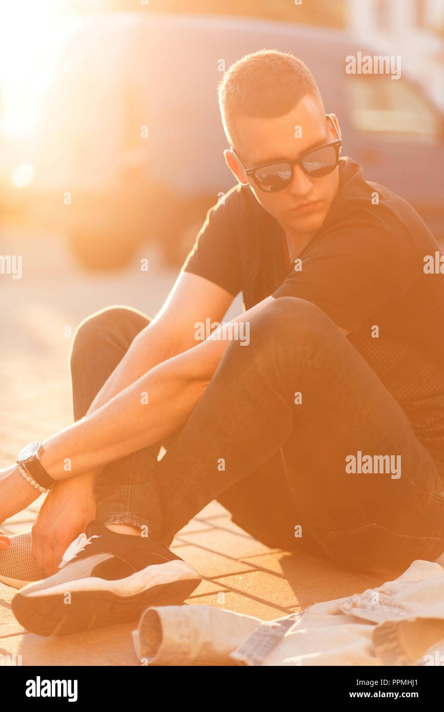 Fashionable handsome young man with a hairstyle in sunglasses and a black T-shirt with jeans with sneakers sits at sunset Stock Photo