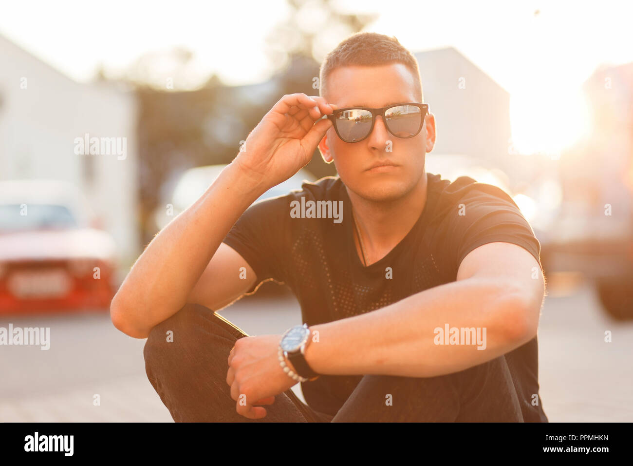 Handsome hipster man with a hairstyle with sunglasses in a black fashion T-shirt is sitting on the street at sunset Stock Photo