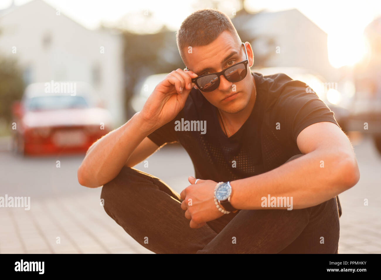 Fashionable handsome young man with sunglasses in a black T-shirt is sitting on the street at sunset Stock Photo