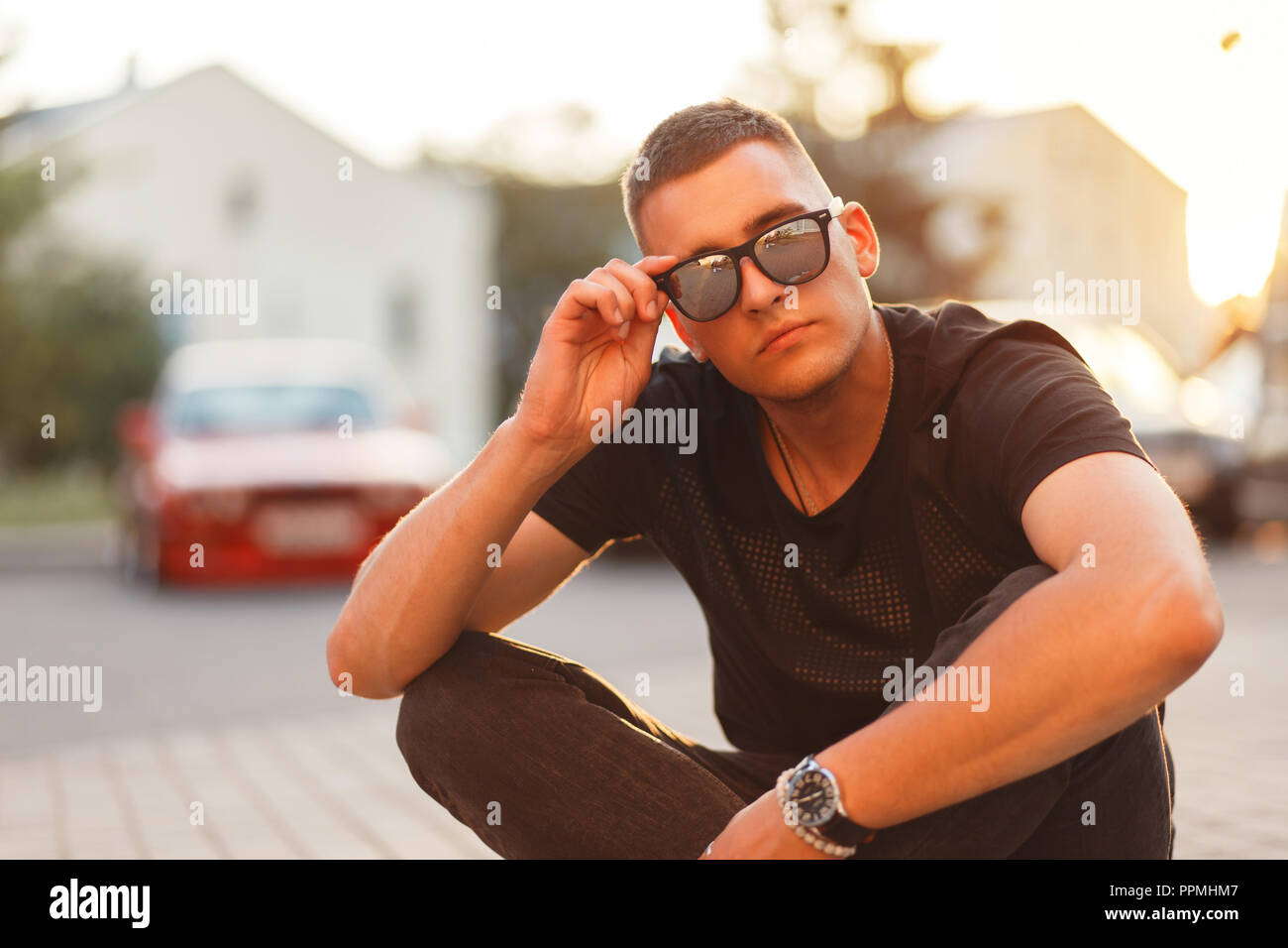 Fashionable handsome young man with black stylish glasses in a black T-shirt is sitting on the street at sunset Stock Photo