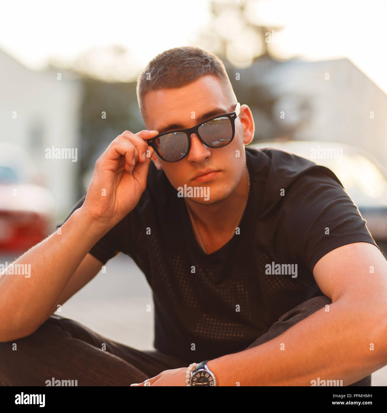 Stylish beautiful young hipster man with a hairstyle in sunglasses and a black T-shirt at sunset sits on the street Stock Photo
