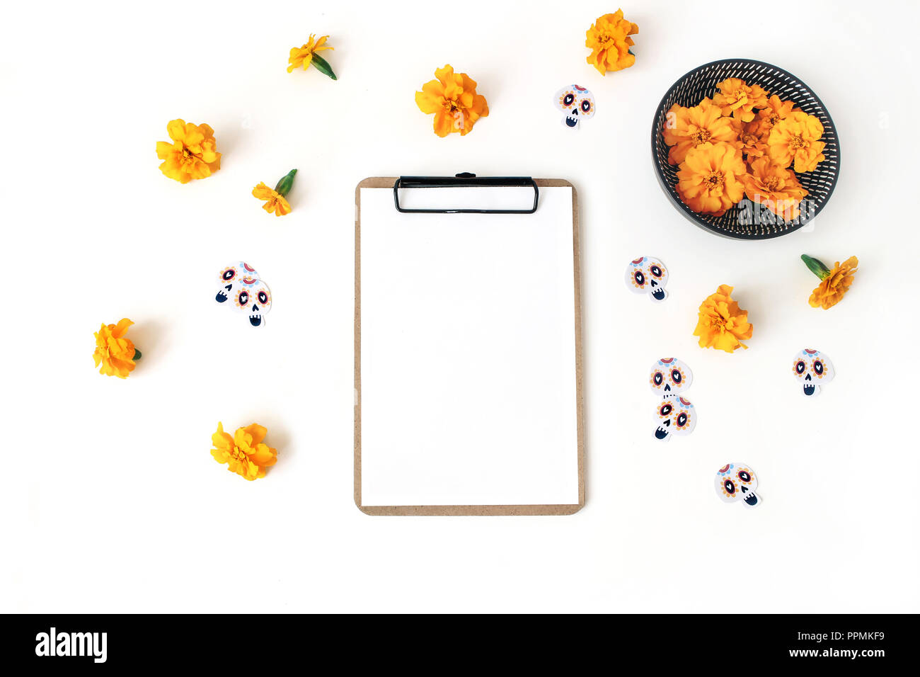 Dia de los Muertos, Mexican Day of the Dead composition. Clipboard, blank paper mockup, orange tagetes, marigold flowers and decorative paper skull stickers. White table background. Flat lay, top view Stock Photo