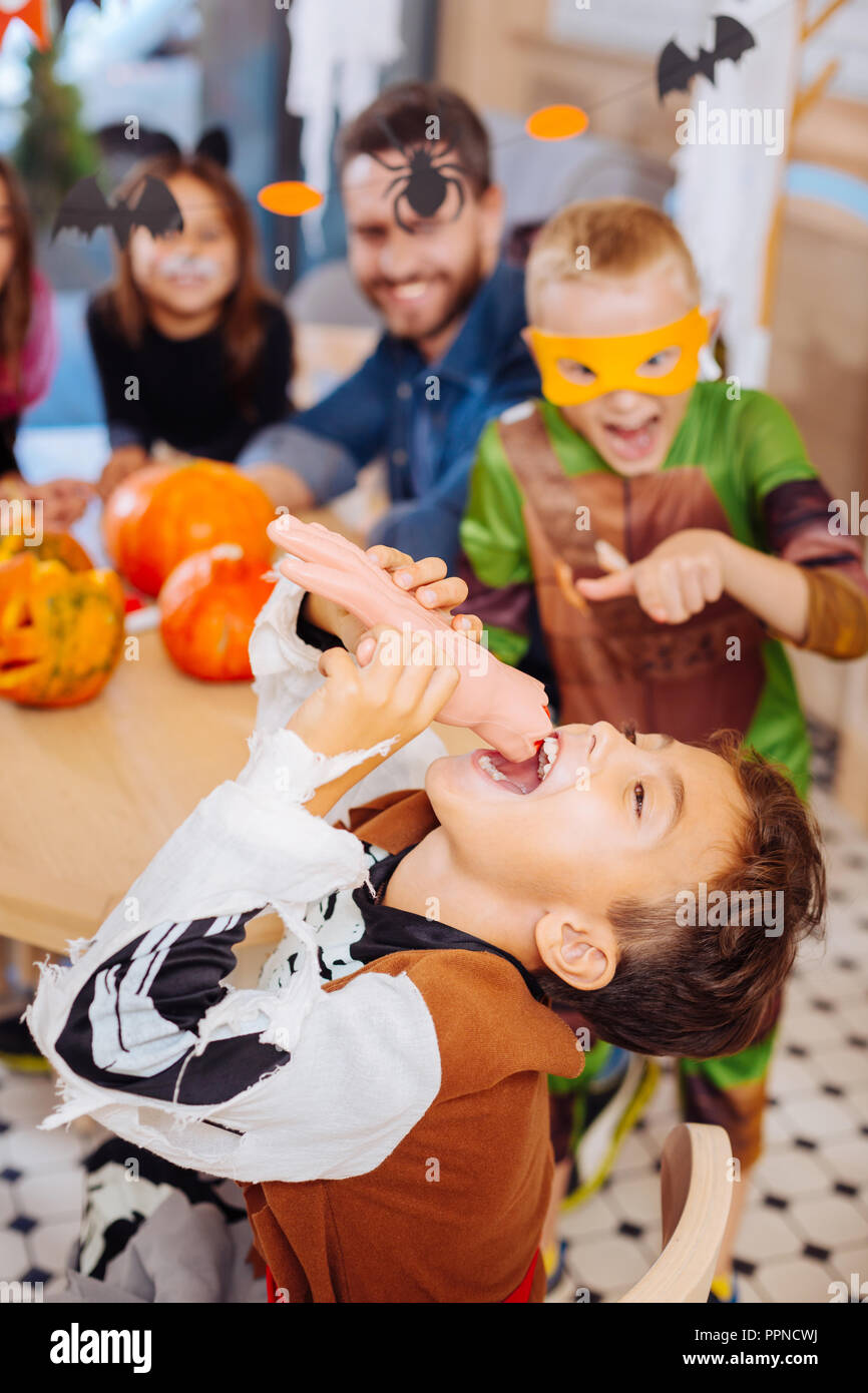 Dark-haired funny boy wearing skeleton Halloween costume eating hand cookie Stock Photo
