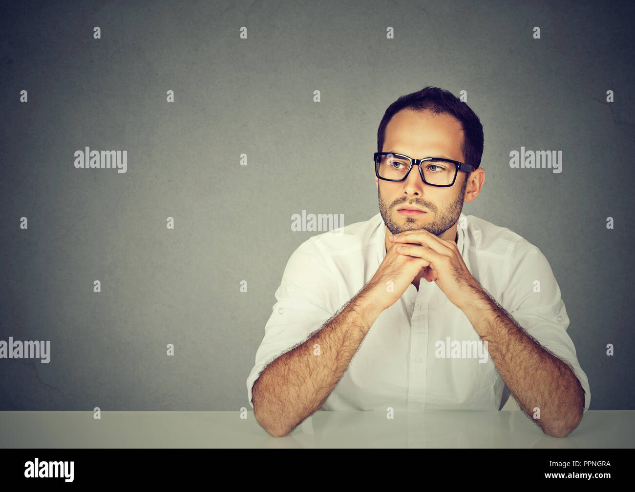Adult sad man in white shirt and glasses sitting at table leaning on hands and thinking on gray background Stock Photo