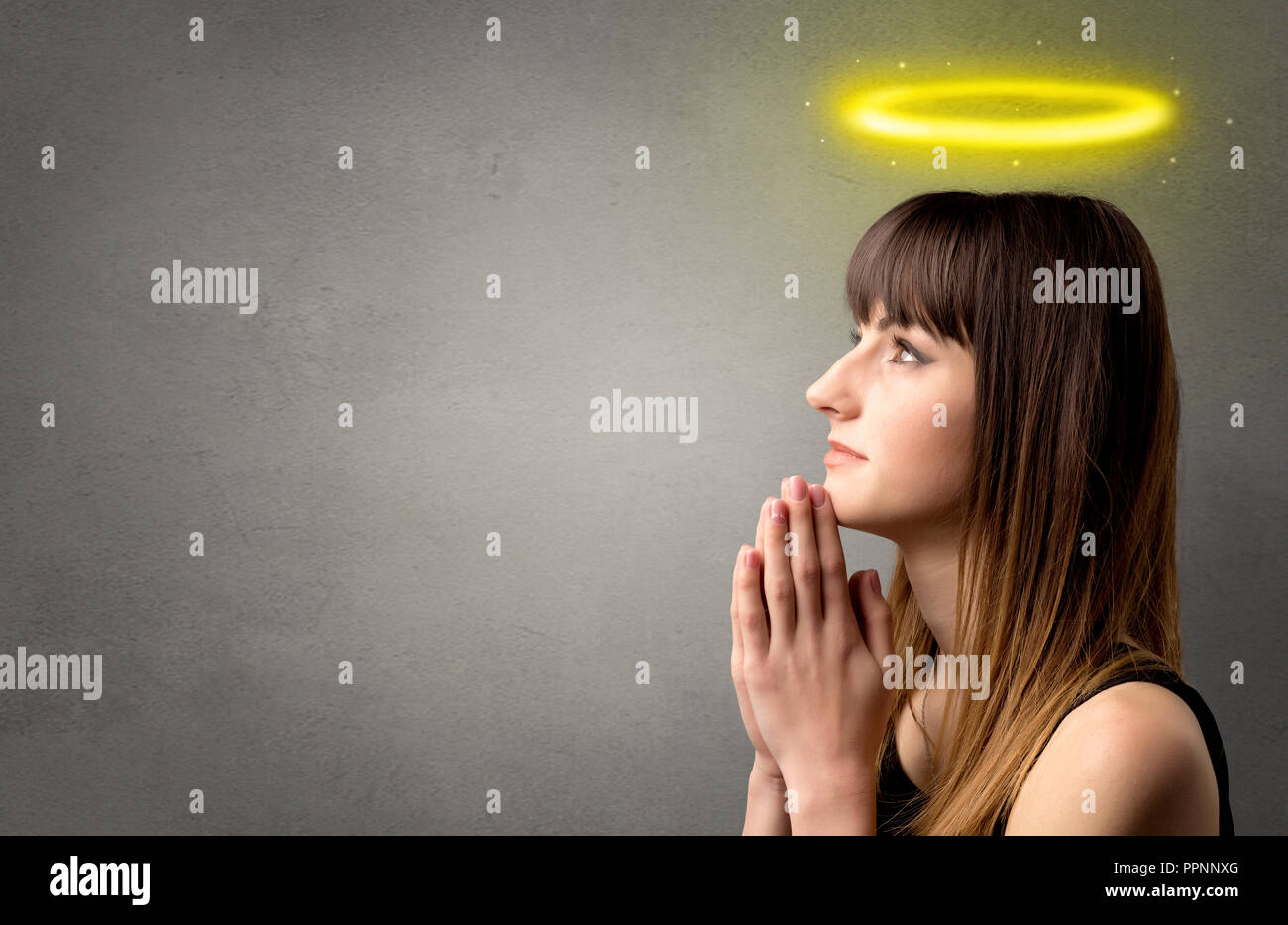Young woman praying on a grey background with a shiny yellow halo above ...
