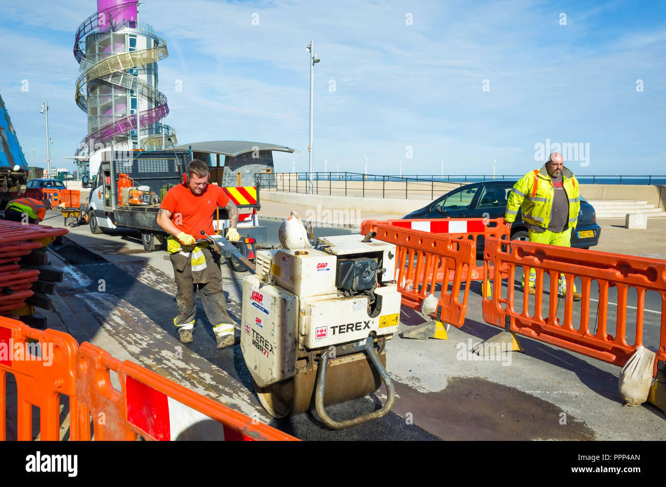 Road repairs on Redcar Seafront with a workman operating a hand controlled power roller compacting tarmac over a refilled cable trench Stock Photo