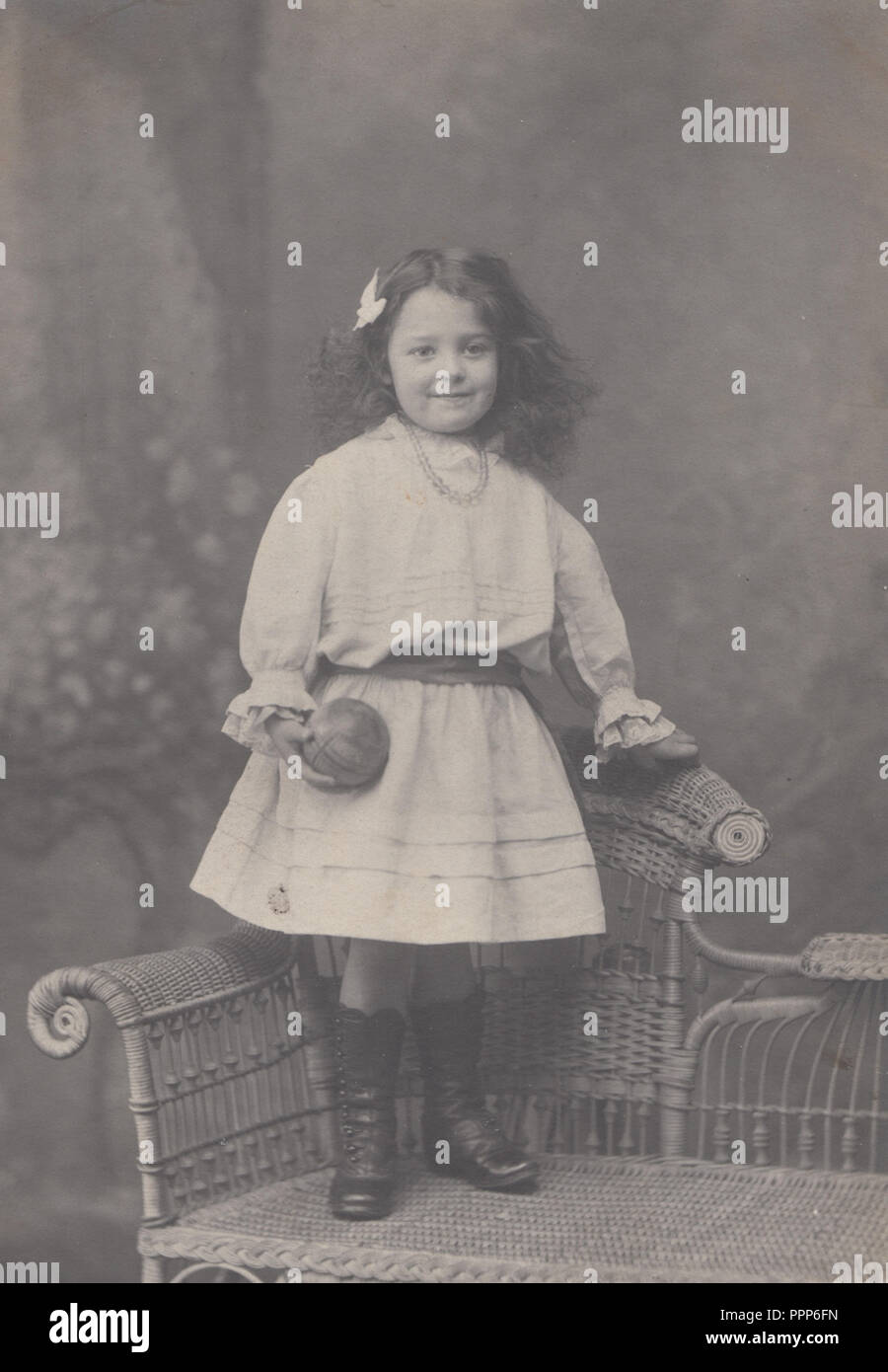 Vintage Southport Studio Photograph of a Young Girl Stood on a Wicker Chair Holding a Small Ball Stock Photo