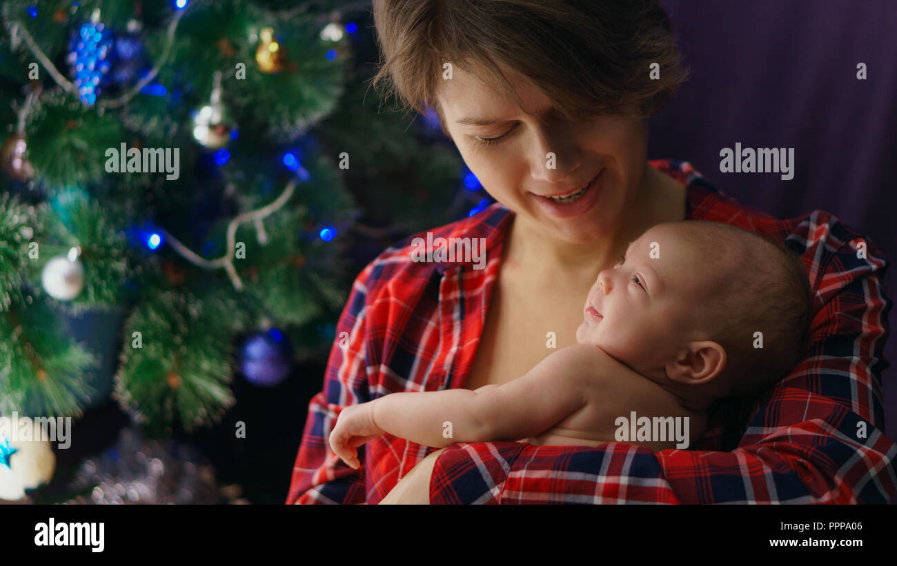 Family celebration. Portrait of young mother and newborn baby. They are smiling. In background is Christmas tree. Happiness of motherhood. Stock Photo