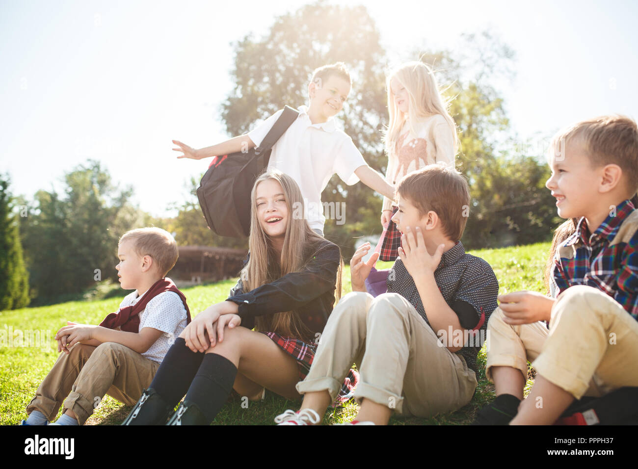 A group of happy smiling children of school and preschool age are sitting on the green grass in the park. The childhood, Kids fashion, school, education, friends, lifestyle, leisure, schoolchildren concept Stock Photo