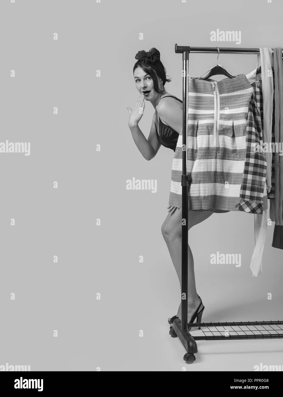 Portrait of beautiful surprised woman in black bikini , standing near hangers with clothes, thinking over what to buy. Black and white. Stock Photo