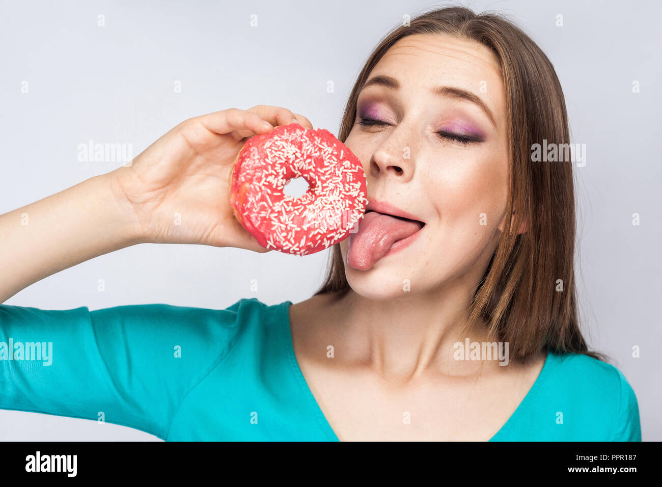 Portrait of pleasure dreaming young beautiful girl in blue blouse standing, holding and showing pink donut, trying to suck with tongue out in grey bac Stock Photo