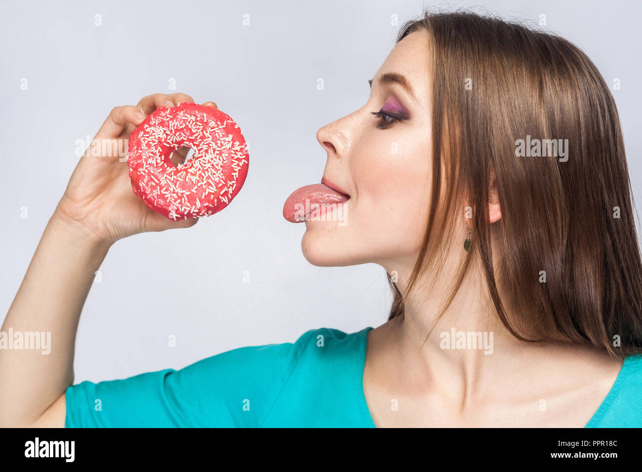 Side view profile of cheerful young beautiful girl in blue blouse standing, holding and showing pink donut, trying to suck with tongue out in grey bac Stock Photo
