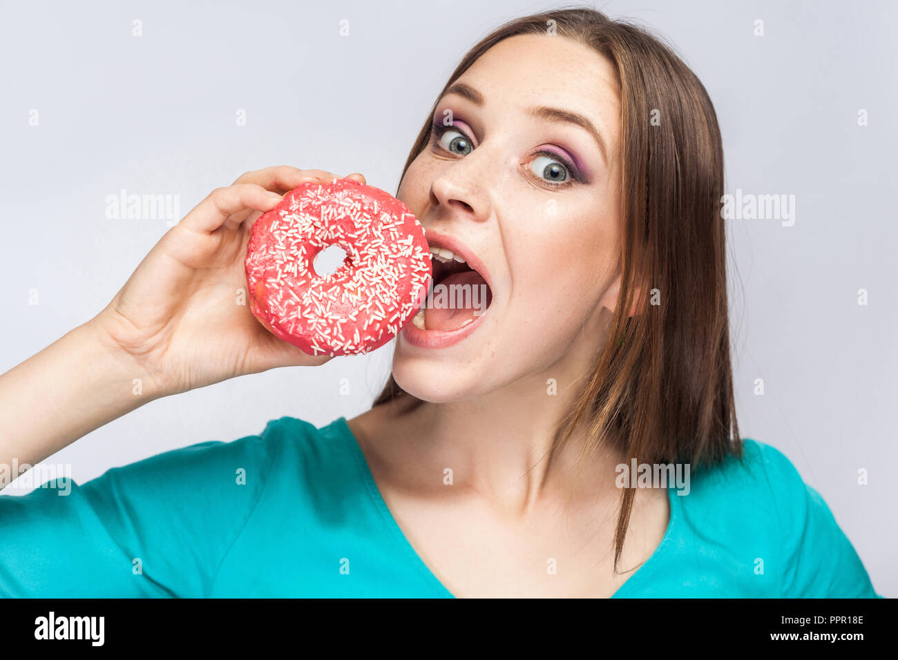 Portrait of hungry young beautiful girl in blue blouse standing, holding and trying to bite big pink donut with open mouth and big eyes in grey backgr Stock Photo