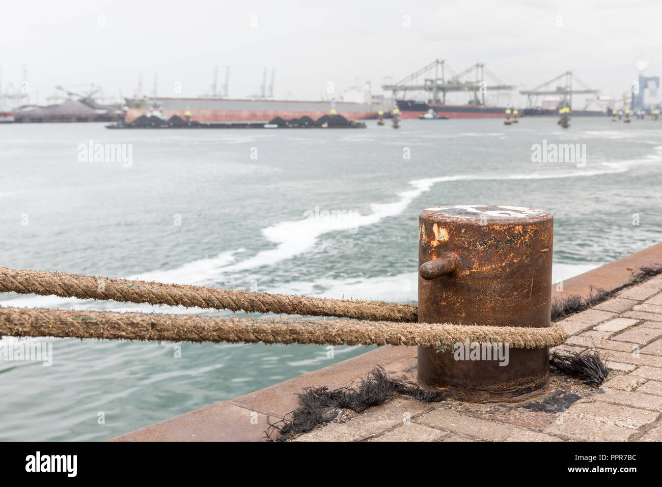 A iron bollard with a tied rope on a quay in the Port of Rotterdam in the Netherlands. In the background, slightly out of focus, is the industrial are Stock Photo