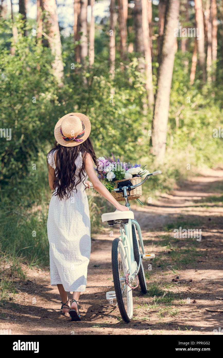 back view of girl in straw hat and white dress walking with bicycle and ...