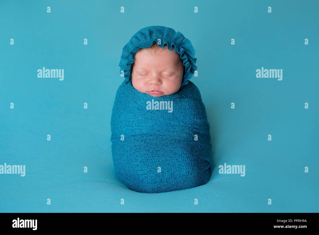 A three week old newborn baby girl wearing a turquoise blue bonnet. She is sleeping upright while swaddled in a stretch wrap. Stock Photo