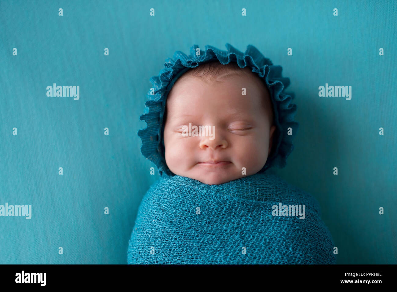 A smiling, three week old, newborn baby girl wearing a bonnet and bundled up in a turquoise blue swaddle. Stock Photo
