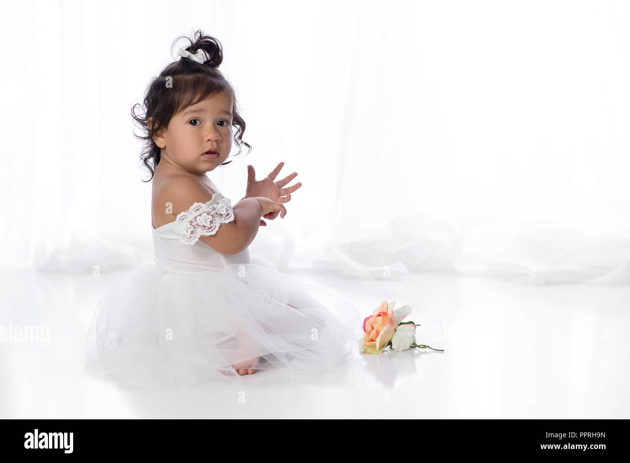A one year old, baby girl sitting on the floor with a flower. She is wearing a white, tulle dress wtih lace sleeves. Stock Photo