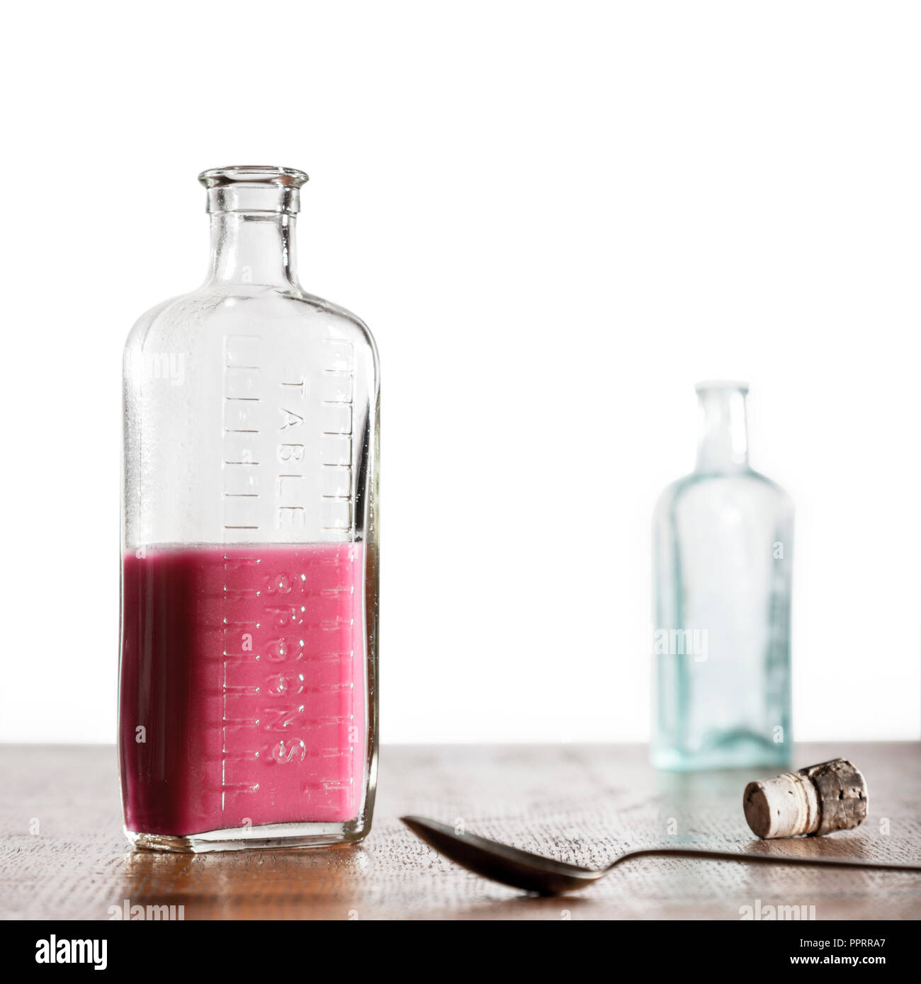 Two bottles. One half full old medicine bottle with a spoon on a wooden surface with a second empty bottle in the background. Stock Photo