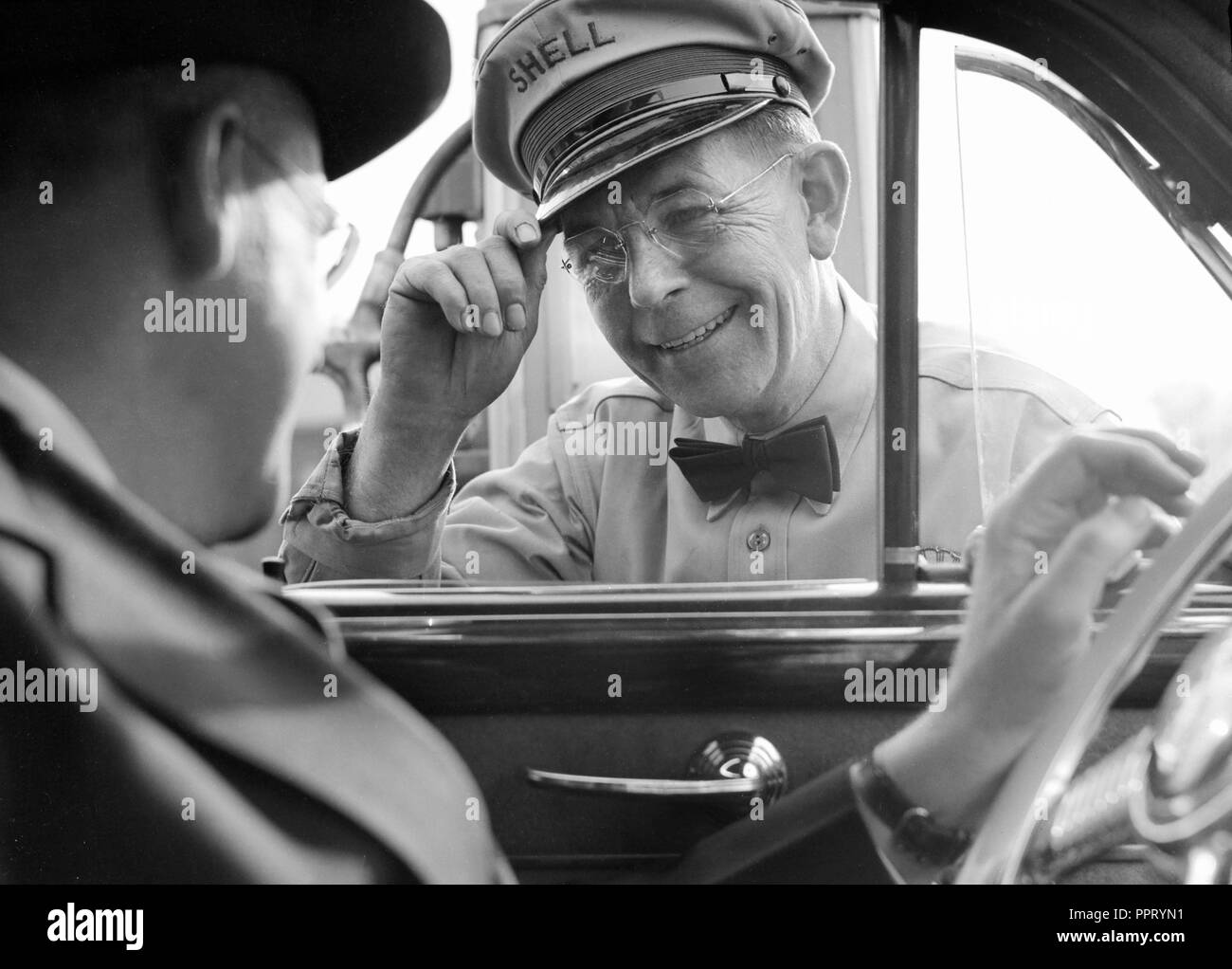 A Shell gas station worker tips his hat to a driver during a service stop, ca. 1940. Stock Photo