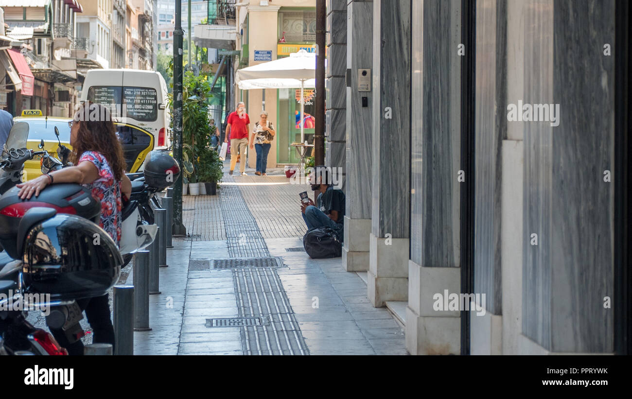 Athens Greece/August 17, 2018: Homeless man with cup and bag sitting on step on streets of Athens with people walking by Stock Photo