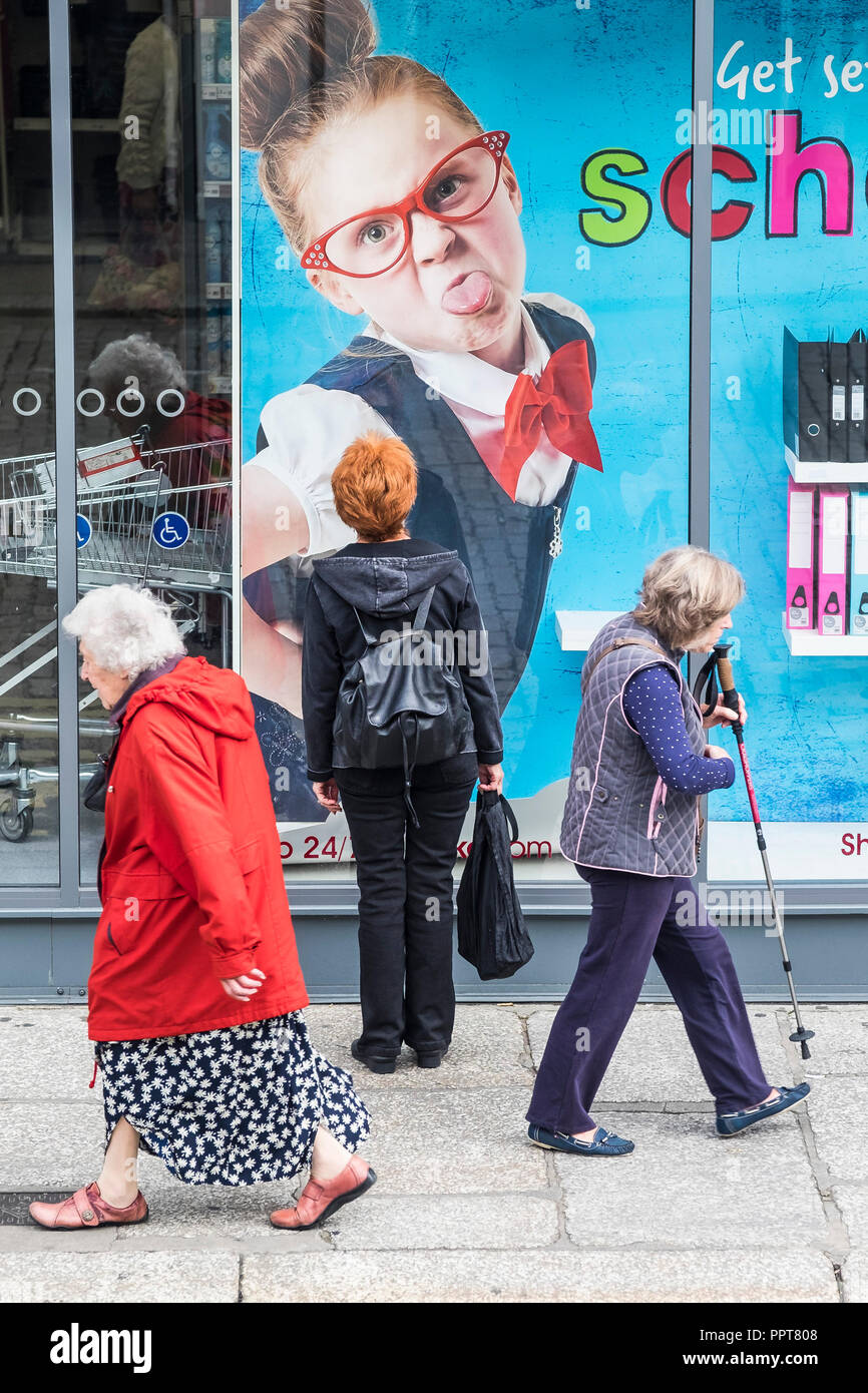 Pedestrians walking on a pavement with one standing and ooking up at a large poster in a shop window in Truro City centre in Cornwall. Stock Photo