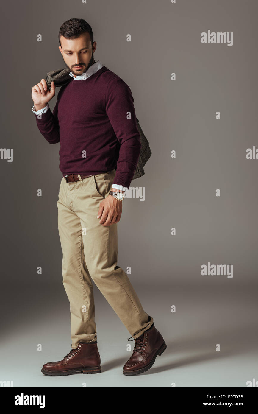 fashionable man posing in beige pants and burgundy sweater with autumn  jacket on shoulder, on grey Stock Photo - Alamy