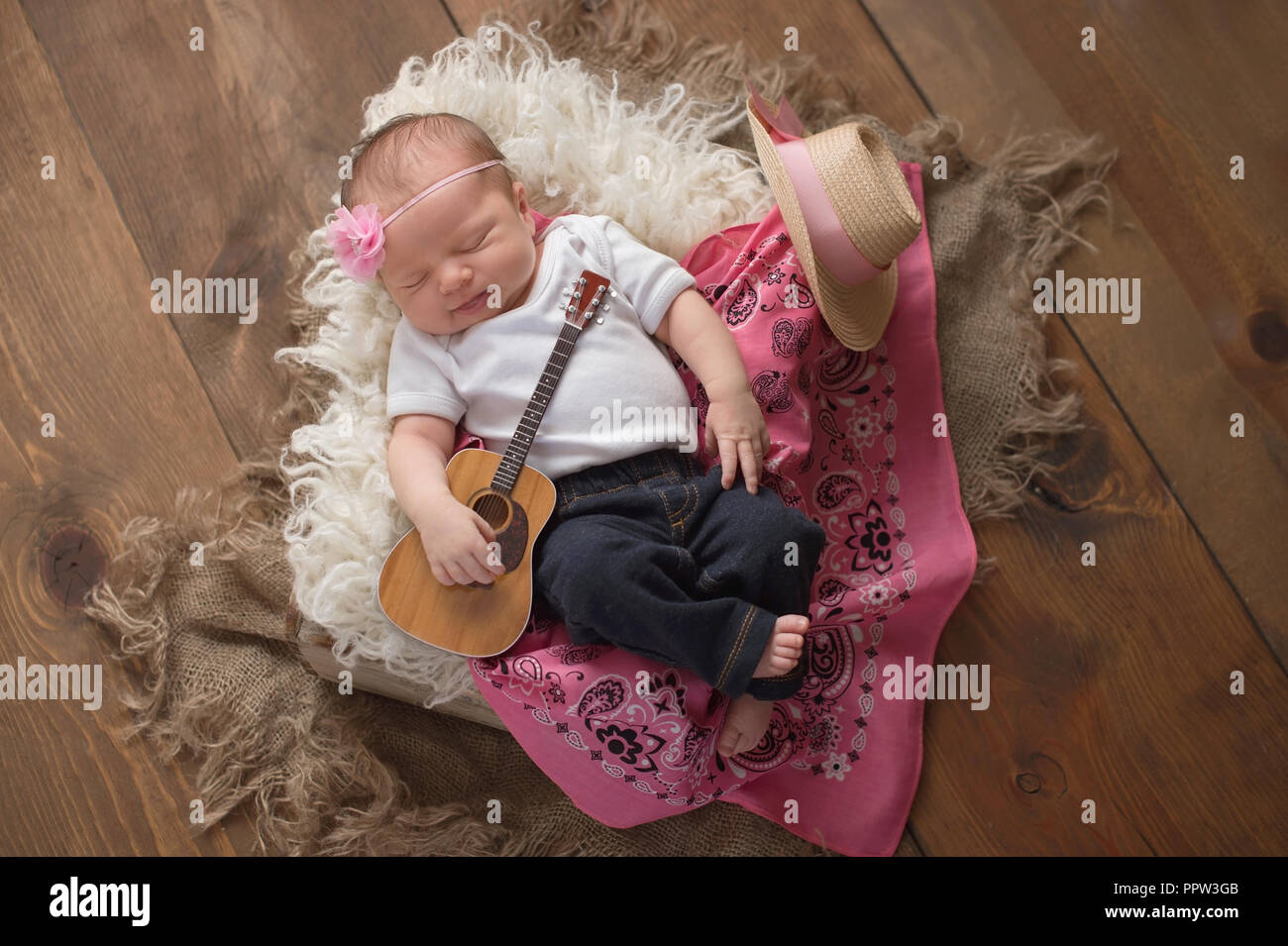 A nine day old, sleeing baby girl wearing jeans and holding a tiny acoustic guitar. She is lying in a wooden crate lined with sheepskin and a pink ban Stock Photo
