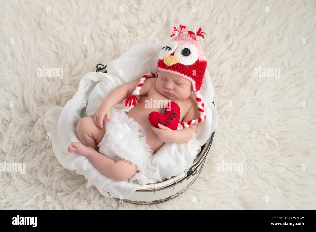 A nine day old, newborn baby girl wearing a pink and red owl hat and holding a heart shaped pillow with the word, 'Love' written on it. Stock Photo